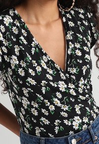 Czarna bluzka w kwiaty z białymi stokrotkami i zielonymi liśćmi. Krótkie rękawy z dekoltem w kształcie litery V, wykonana z miękkiego materiału.
