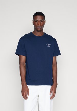 Hombre que viste una camiseta de Tommy Jeans azul marino y pantalones blancos, de pie frente a un fondo claro y liso, mirando hacia adelante.