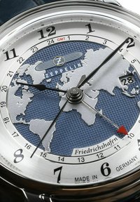 Watch features a blue textured dial with a world map, silver hands, and a date display. Stainless steel case with black accents.