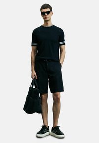 Zwart T-shirt met korte mouwen en witte strepen, zwarte shorts en zwarte sneakers met een witte zool. Houdt een zwarte tas met meerdere vakken vast.