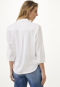 Camicia bianca con colletto a bottoni, maniche a tre quarti e orlo curvo. Tessuto liscio, senza motivi visibili, mostrata da dietro.