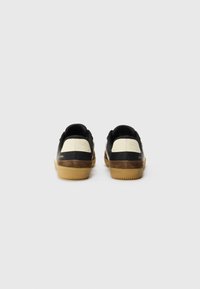 Black sneakers with a textured design, cream accents on the heel, and a rubber gum sole, featuring minimal branding on the side.