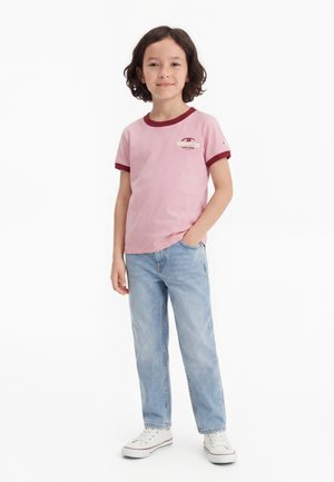 Enfant aux cheveux foncés mi-longs, portant un t-shirt rose Champion avec des finitions rouges, un jean bleu clair et des baskets blanches, debout, une main dans la poche.