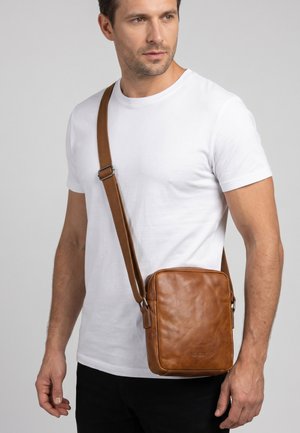 Mężczyzna w prostym białym t-shircie i czarnych spodniach z brązową skórzaną torbą na ramię na szarym tle, patrzy w prawo.