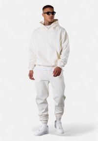 Weißer Baumwoll-Hoodie und Sweatpants-Set mit einer Fronttasche, gerippten Bündchen und kombiniert mit weißen Sneakers. Accessoire: schwarze Sonnenbrille.