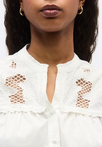 Γυναίκα που φοράει λευκό πουκάμισο με διακοσμητικά λουλουδάτα ανοίγματα δαντέλας στους ώμους και μικρά κουμπιά μπροστά, συνδυασμένο με χρυσούς κρίκους σκουλαρίκια.
