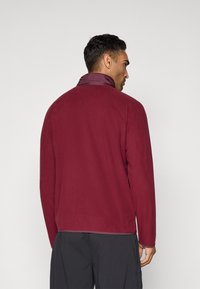 Man facing away wearing a maroon fleece jacket with a high collar and black pants against a plain gray background.