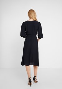 Musta, polvimittainen mekko, jossa on pullottavat hihat ja istuva vyötärö. Kasaava kangas tarjoaa sileän tekstuurin, jota korostavat korkeakorkoiset sandaalit.