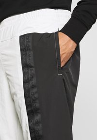 Černé a bílé sportovní kalhoty s elastickým pasem, postranními kapsami a černým pruhovaným logem Adidas podél vnějšího švu. Hladká textura.