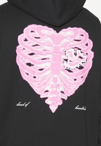 Vaaleanpunainen sydämen muotoinen kylkiluu mustassa hupparissa, jonka takana on valkoiset tekstit "sydäntuskan saari" kuvion alapuolella.