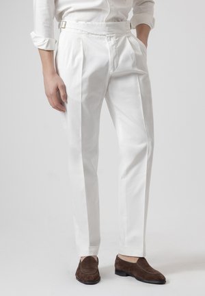 Persona con pantalones blancos plisados y cinturón, y mocasines de ante marrones, de pie con una mano en el bolsillo.