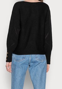 Der schwarze Strickpullover hat einen weiten, offenen Rücken, eine gerippte Textur und Knopfdetails an den Ärmeln, kombiniert mit hellblauen Jeans.