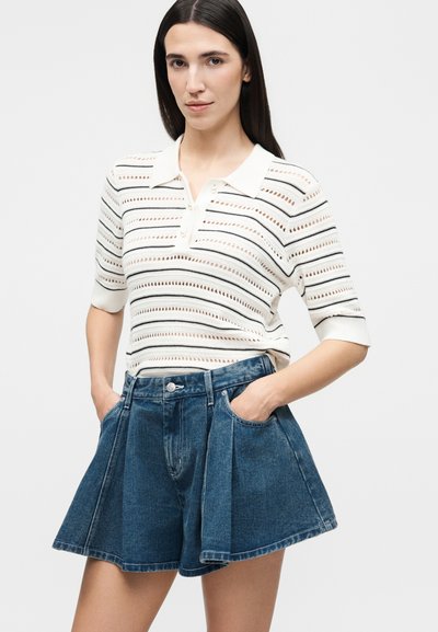 Νεαρή γυναίκα φοράει λευκό ριγέ πόλο με κοντά μανίκια, το οποίο έχει μπει μέσα σε ψηλόμεσες, πλισέ κοντές τζιν βερμούδες, με τα χέρια στις τσέπες.