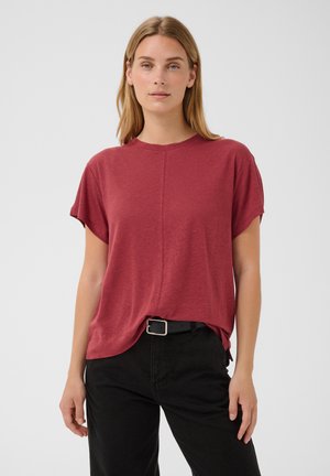 Jeune femme aux cheveux blonds lisses portant un t-shirt rouge à manches courtes rentré dans un pantalon noir avec une ceinture noire, debout devant un fond uni.