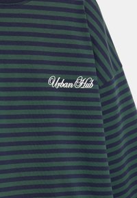 Raidallinen t-paita vihreillä ja laivastonsinisillä väreillä, pehmeällä tekstuurilla ja pyöreällä kaula-aukolla. "UrbanHub"-logo on kirjailtu valkoisella.