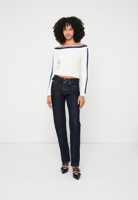 Bijeli rebrasti džemper s otvorenim ramenima s plavim naglascima, u kombinaciji s tamnim trapericama i crnim cipelama na visoku petu s ukrasima.