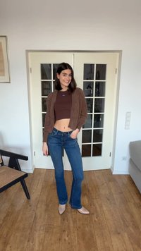 Νεαρή γυναίκα που χαμογελά, στέκεται μέσα σε εσωτερικό χώρο, φορώντας καφέ κοντό τοπ, αντιστοιχισμένο ζακέτα, μπλε τζιν και μπεζ παπούτσια, με το χέρι στην τσέπη.