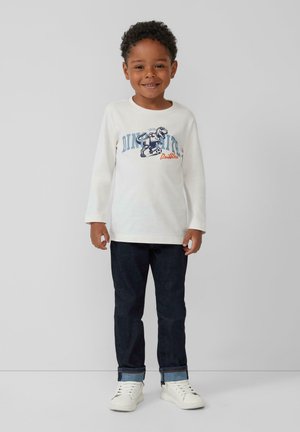 Weißes langärmliges T-Shirt mit einem Grafikdruck eines Dinosauriers, kombiniert mit dunklen Jeans und weißen Sneakern.
