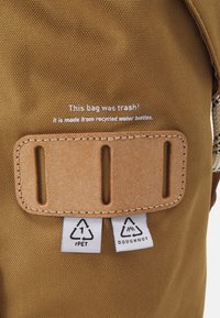 Sac à dos en tissu recyclé de couleur marron avec une texture lisse, comprenant un accent en cuir et des étiquettes indiquant des matériaux écologiques.