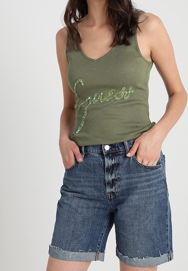 Kvindelig iført en olivengrøn ærmeløs top med "Sweet" i rhinsten og opsmøgede denimshorts, hænderne i lommerne, mod en ensfarvet baggrund.