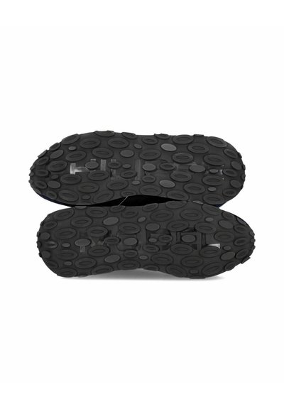 Chaussures en caoutchouc noires montrées par le dessous, avec une semelle texturée présentant des grips en forme d'ovale pour une adhérence optimale.