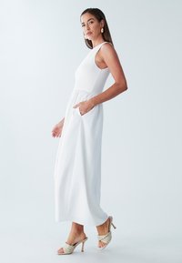 Balta ilga suknelė su tekstūruotu korpusu, be rankovių ir su laisvai krentančia suknele. Turi dvi šonines kišenes ir derinama su austa avalyne tipo mule.