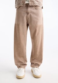Jeans de pierna ancha color marrón claro con una textura desgastada. Cuenta con bolsillos clásicos y un cordón en los puños. Combinados con zapatillas blancas.