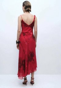 Robe florale rouge à bretelles spaghetti, dos en V, tissu fluide et ourlet jusqu'aux chevilles. Talons aiguilles complètent la tenue.