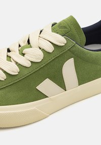 Green suede sneaker featuring cream laces, a textured rubber sole, and a prominent white logo accent on the side.