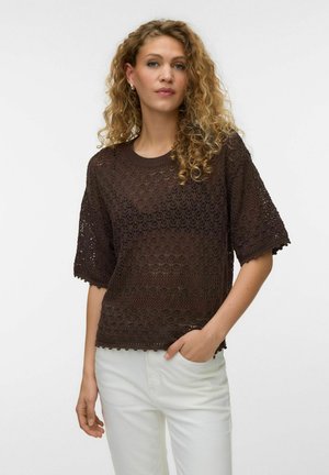 Kvinde med krøllet hår iført en mørkebrun kortærmet hæklet top og hvide bukser, med den ene hånd i lommen.