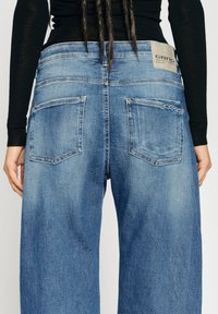 Korkeavyötäröiset, löysät siniset farkut, joissa on haalistunut pinta. Varustettu kahdella takataskulla ja vyötäröllä ommellulla tarralla.