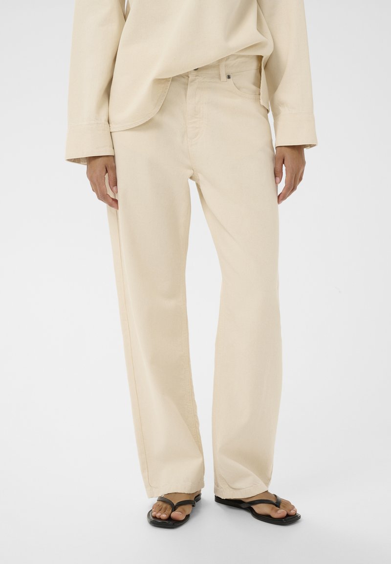 Persoon die losse beige broek, bijpassend beige shirt met lange mouwen en zwarte slippers met vierkante neus draagt, staand op een witte achtergrond.