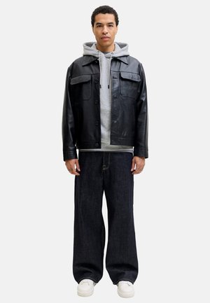 Mand iført sort læderjakke over grå hættetrøje, løstsiddende mørke jeans og hvide sneakers, stående foran ensfarvet baggrund.