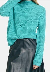 Suéter de punto en color verde azulado con cuello alto, que presenta patrones de olas texturizados y puños acanalados, combinado con una falda de cuero negra.