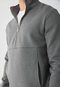 Hanorac gri cu fermoar și guler înalt, confecționat dintr-un material moale. Dispune de buzunare laterale și un design de cusătură în contrast.