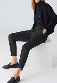 Personne assise sur un bloc blanc portant une veste noire zippée, un haut noir, un pantalon gris foncé retroussé et des mocassins noirs, les mains dans les poches.