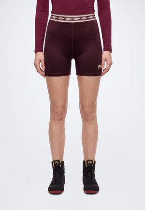 Le modèle se tient face à l’avant, vêtu d’un short de sport bordeaux avec une taille à motifs et de chaussures montantes noires, sur un fond blanc uni.