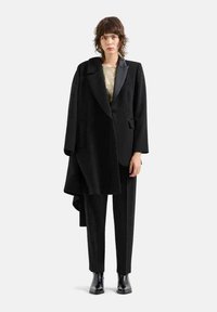 Cappotto nero su misura con vestibilità oversize, design doppiopetto e rever in raso; indossato su una maglietta leggera e abbinato a pantaloni neri e stivali.