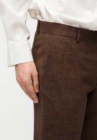 Main reposant à côté d'un pantalon habillé marron texturé porté avec une chemise blanche à manches longues avec larges poignets.