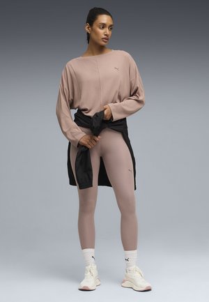 Femme vêtue d'un haut mauve à manches longues et de leggings avec des baskets blanches, attachant une veste noire autour de la taille sur un fond dégradé gris.
