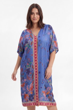 Femme aux cheveux bruns mi-longs portant une robe bleue ample longueur genou avec des motifs floraux colorés et une bande verticale rouge à motif.