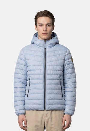 Giovane uomo che indossa una giacca trapuntata azzurro chiaro con cappuccio, tasche con cerniera e chiusura con cerniera, in piedi su sfondo bianco semplice.