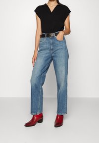 Schwarzes kurzärmeliges Poloshirt, hoch sitzende hellblaue Jeans und rote Lederstiefeletten mit niedrigem Absatz. Lässiger Schnitt, minimale Details.