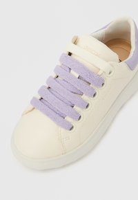 Krém színű sportcipő puha levendula színű fűzőkkel, sima bőrtapintással, és egy finom logóval a nyelvén, kerek orral és gumitalppal.