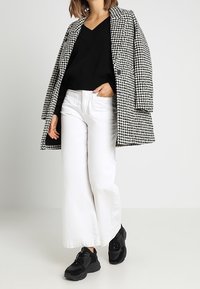 Abrigo de cuadro houndstooth en blanco y negro, suéter negro, pantalones anchos blancos y zapatillas gruesas negras. Texturas suaves y un corte holgado.