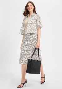 Lined geometric-patterned dress with a cropped jacket, both in cream and black. Model holds a textured black handbag and wears strappy heels.