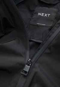 Veste imperméable noire avec fermeture à glissière et texture lisse. Comprend une étiquette indiquant "NEXT" et "MADE IN MYANMAR" à l'intérieur.