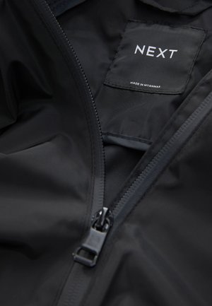 Veste imperméable noire avec fermeture à glissière et texture lisse. Comprend une étiquette indiquant "NEXT" et "MADE IN MYANMAR" à l'intérieur.