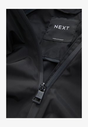 Chaqueta impermeable negra con cierre de cremallera y textura suave. Cuenta con una etiqueta que dice "NEXT" y "HECHO EN MYANMAR" en el interior.