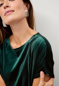 Grøn fløjlstop med en afslappet pasform, der har rund halsudskæring og samlede ærmedetaljer. Metaløreringe giver et kontrasterende element.